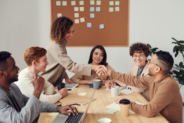 Réunion de six employés autour d'une table. Une des personne sert la main au nouvel arrivant, en signe de bienvenue dans l'entreprise.